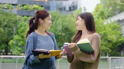 Two young Asian female students enjoy a lively study session outside their university.