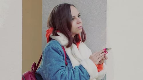 Young Beautiful Woman in Warm Denim Jacket Using Smartphone Leaning Against Wall on Street