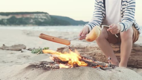 Boy fries sausage and bread on strong fire, sitting on sand by sea