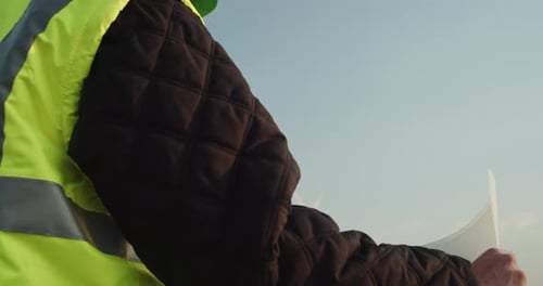 Engineer wearing a reflective vest and helmet with design plans stands in front of a wind turbine fa