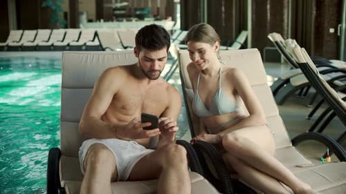 Couple lounges by indoor pool looking at phone