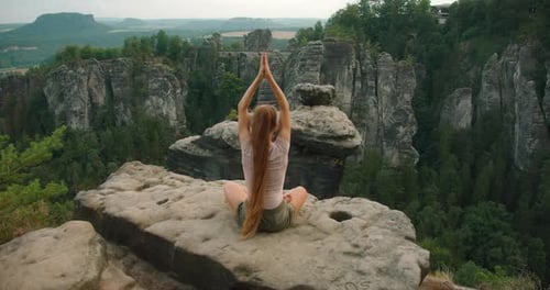 Woman with long hair raising arms in a yoga pose on a rocky cliff with a picturesque forest and rock