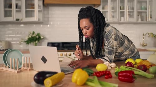 Woman Eats Vegetables While Using Laptop in Kitchen