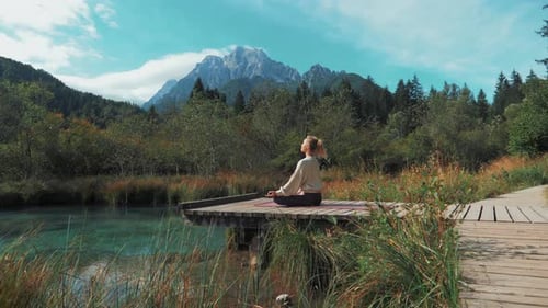 A beautiful shot is moving towards a young woman meditating on a wooden path surrounded by nature.