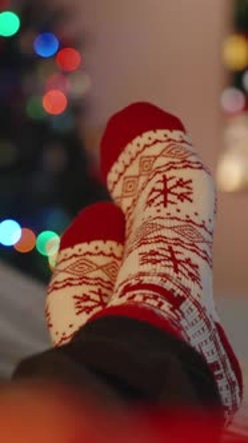 Vertical video. Feet in Festive Christmas Socks Propped Up, Relaxing by a Lit Christmas Tree and TV.