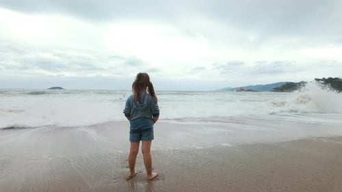 Child Stands on a Sandy Beach Looking Out to Sea A Wave Hits the Shore and the Girl Runs Away