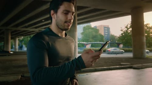 Athletic Muscular Young Man in Sports Outfit Jogging in a Street. He's Using His Smartphone on the