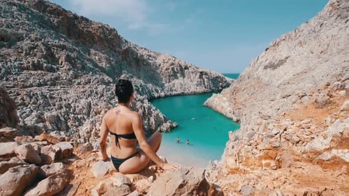 A woman sitting on the ledge, overseeing a turquoise bay.