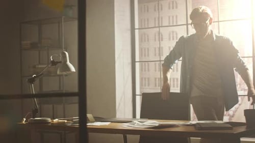 Creative Young Designer Walks into His Studio Office, Sits at His Desk, Opens Laptop and Starts Wor