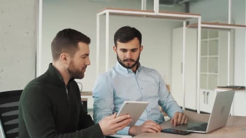 Two adult men are sitting in a stylish office with a laptop. One man is showing his completed work o