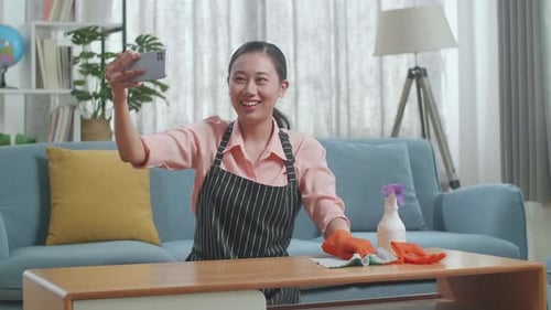 Asian Female Housekeeper Having Video Call On Smartphone While Cleaning The Table
