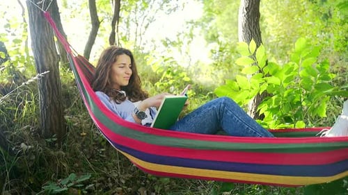 Encantadora jovencita de cabello oscuro acostada en una colorida hamaca en el parque de verano y leyendo un libro nuevo.
Co