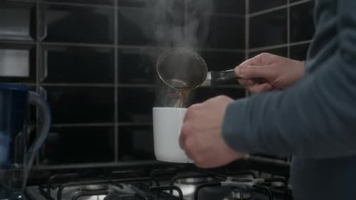 Close Up View of Pouring Coffee From Chromed Metal Cezve Into White Mug Against Dark Kitchen