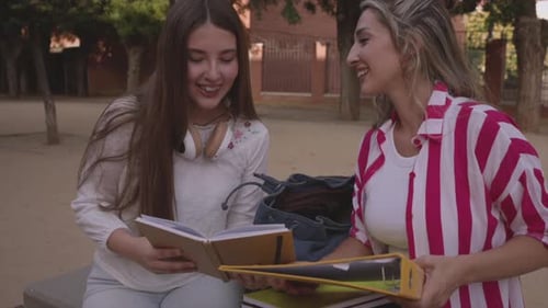 Two female students are sitting on a bench in a campus park, sharing notes and studying together, en