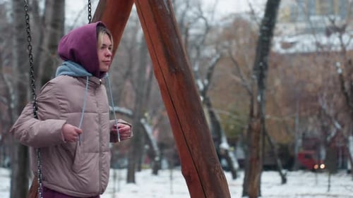 Calm Young Woman Swinging in Winter Park Holding Chains with Hoodie