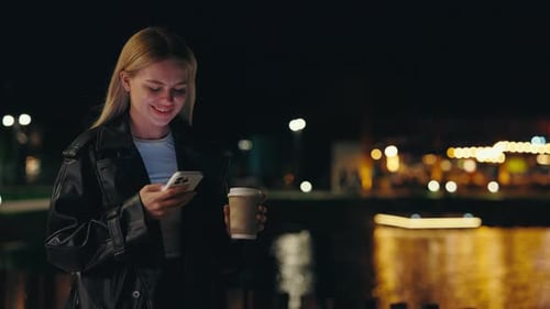 Cute Young Business Lady Using Smartphone Standing on the Night Street with Cup of Coffee