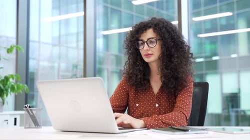 Confident businesswoman works on laptop while sitting at desk at workplace in modern glass business