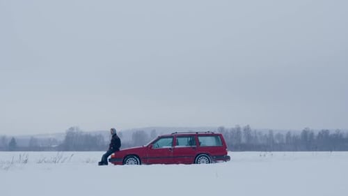 Frustrated man sit on hood of red broken down car in white winter snow landscape