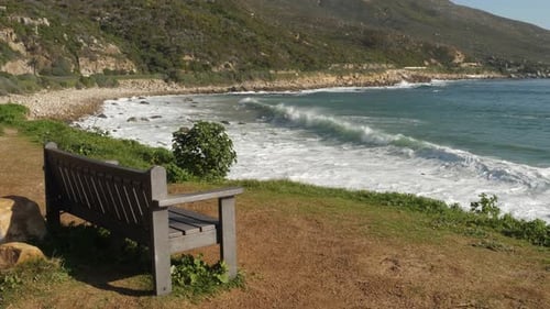 Bench overlooks surf waves breaking onto small rocky beach in Cape town