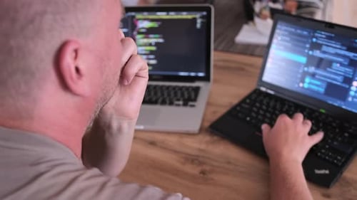 Rückansicht, Nahaufnahme, IT-Spezialist tippt zu Hause auf einem Laptop, umgeben von Monitoren