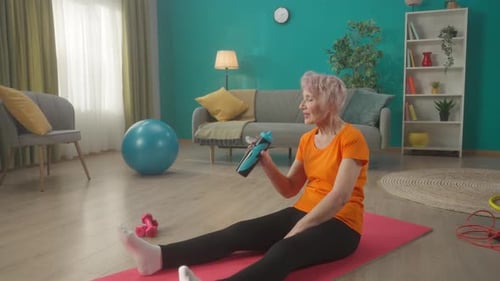 An Elderly Woman Tired After a Workout Sits on a Gym Mat in the Living Room The Woman Runs Her Hand