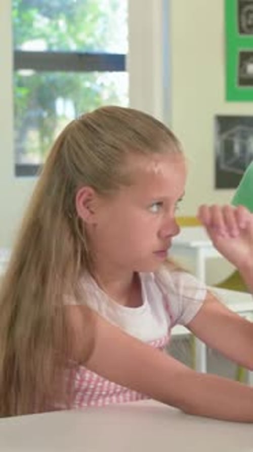 Vertical video: Reacting to peer entering frame, girl wearing pinafore in classroom tapping tablet