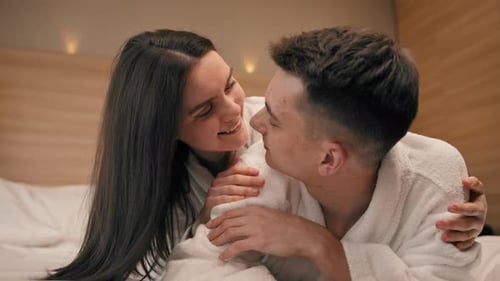 Porträt eines jungen Paares in den Flitterwochen im Hotelzimmer, das in weißen Gewändern auf dem Bett liegt und glückliche Liebhaber küsst
