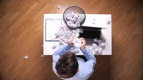 Man at Cluttered Office Desk Crumpling Paper in Frustration Battling Writer's Block and Exhaustion