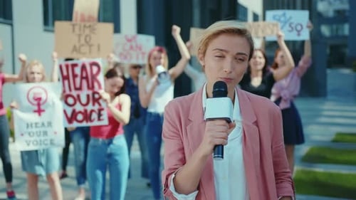 Female Reporter Speaks to Crowd Holding Protest Signs