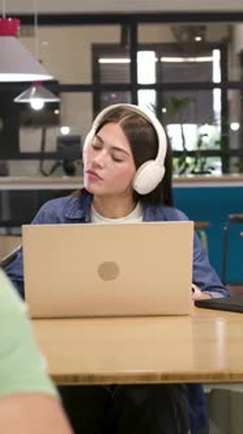 Vertical video: Listening, denim-clad woman taking notes with pencil at communal table with laptop