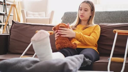 Menina com perna quebrada sentada no sofá Criança triste abraçando seu ursinho de pelúcia se recuperando