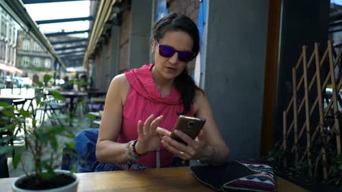 Pretty young woman in sunglasses uses smartphone at cafe table outdoors