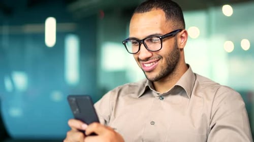 Happy handsome male employee in glasses using mobile phone while sitting in modern glass office