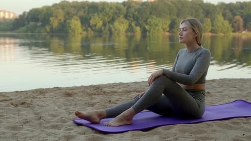 Slender Young Woman Sit on Purple Sport Mat on Sandy Shore of River and Look Into Distance