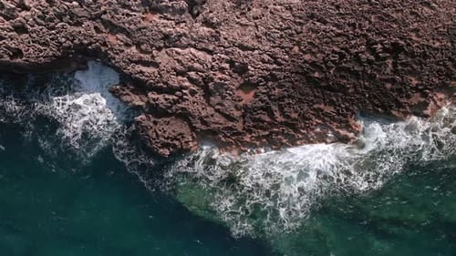 4K aerial drone shot gliding left to right over rugged coastal rocks as ocean waves crash below. A c