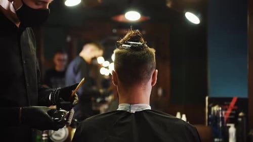 Young bearded man sitting and getting haircut in barber shop by worker that uses scissors