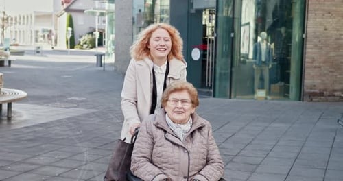 Smiling Woman Pushes Senior in Wheelchair in City