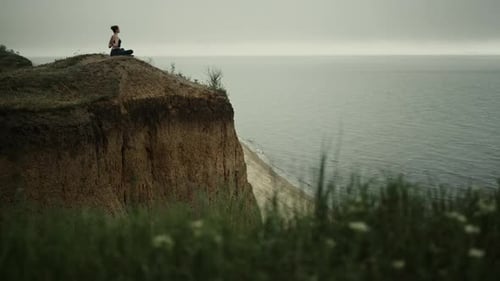 Junge Frau praktiziert Yoga-Lotusposition auf einem grünen Hügel mit Blick auf den ruhigen Ozean