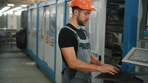 Young worker in uniform stands in the factory and typing on the laptop