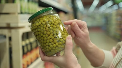 Woman reading label on glass jar of canned green peas before buying in supermarket