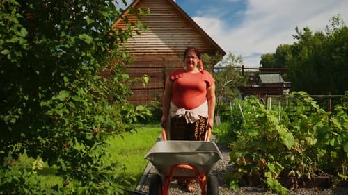 A Woman Pushing a Wheelbarrow in a Vibrant Garden Surrounded By Lush Greenery and a Rustic Wooden