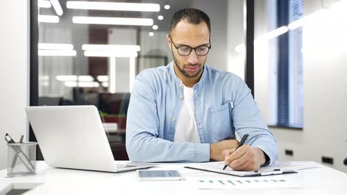 Busy african american financier is doing paperwork while sitting at desk in business office.