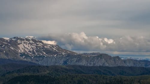 Zeitraffer mit Panoramablick auf die Berge mit schneebedeckten Gipfeln, dichten Wäldern und dramatisch bewölktem Himmel i