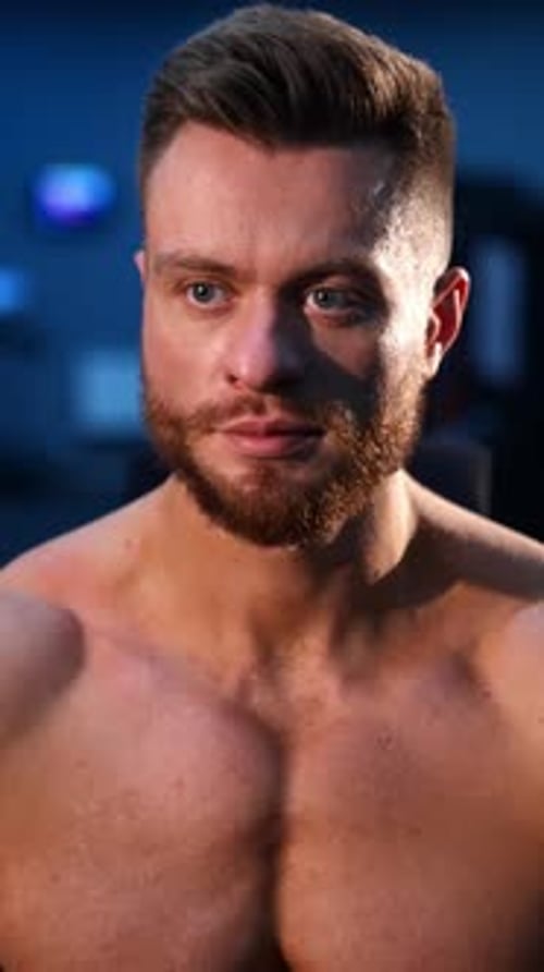 Portrait of bodybuilder indoors. Young bearded sportsman with athletic body sitting in gym.