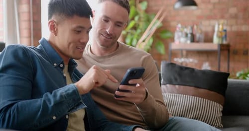 Two Mid Adult Males at Home Sitting on a Sofa looking at a Smartphone Together
