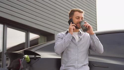 Hübscher Mann telefoniert und Kaffee trinkt, während er ein Luxus-Elektroauto am Bahnhof auflädt