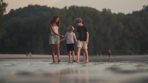 Children Holding Hands Strolling Along Beach With Dense Greenery Behind