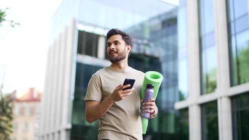 Happy sporty man walking on city street using browsing mobile phone. Joyful young adult male athlete