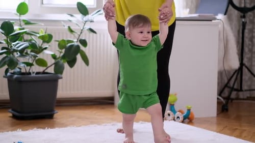 Caring Mother Teaching Little Son to Walk on White Rug Holding Hands