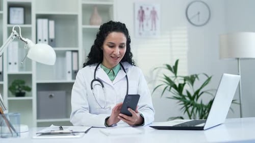 Happy female doctor in white coat using smartphone, typing message in modern hospital clinic. A smil